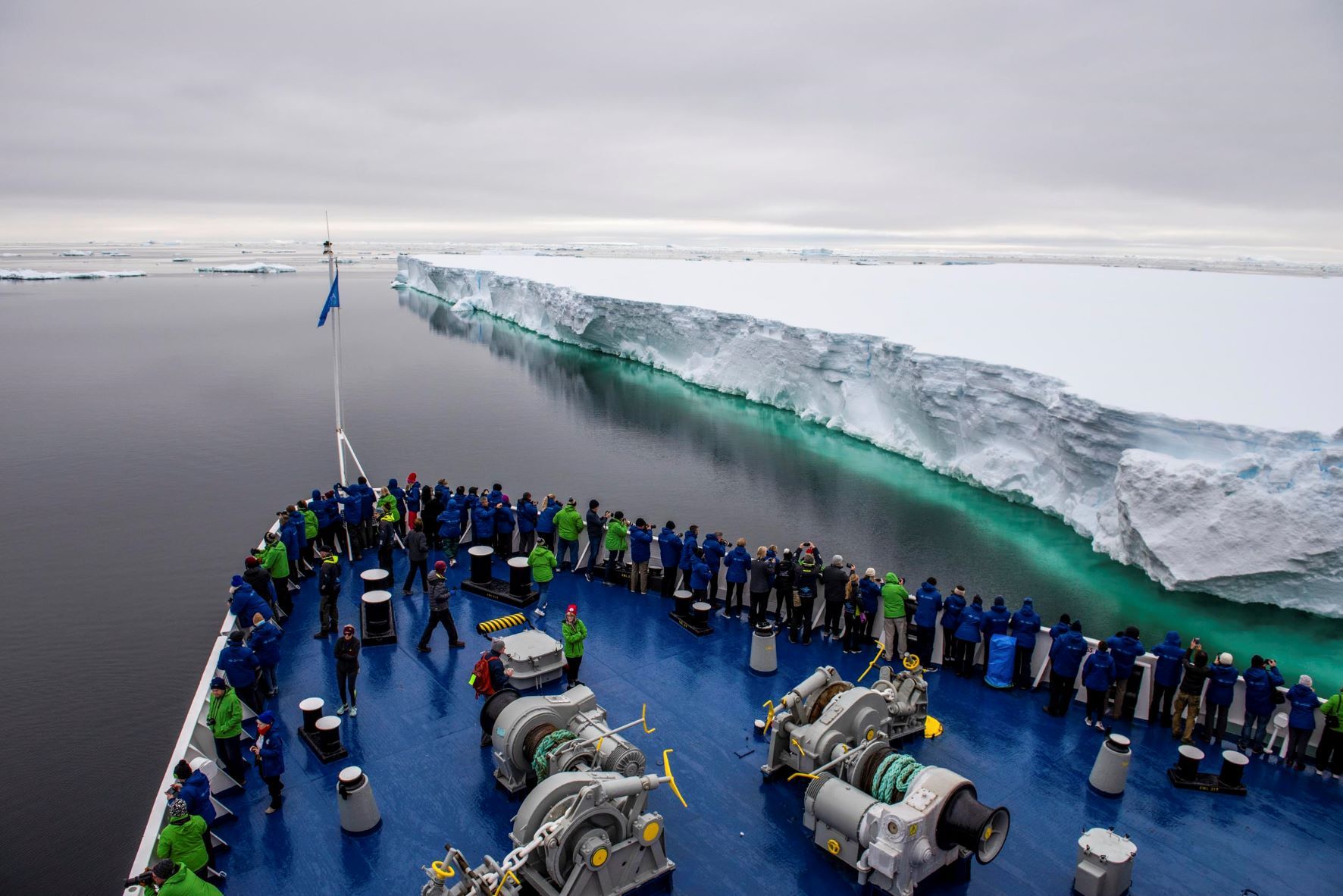 Expedition cruise passengers lining the bow deck to photograph a massive tabular iceberg in Antarctica after crossing the Drake Passage.