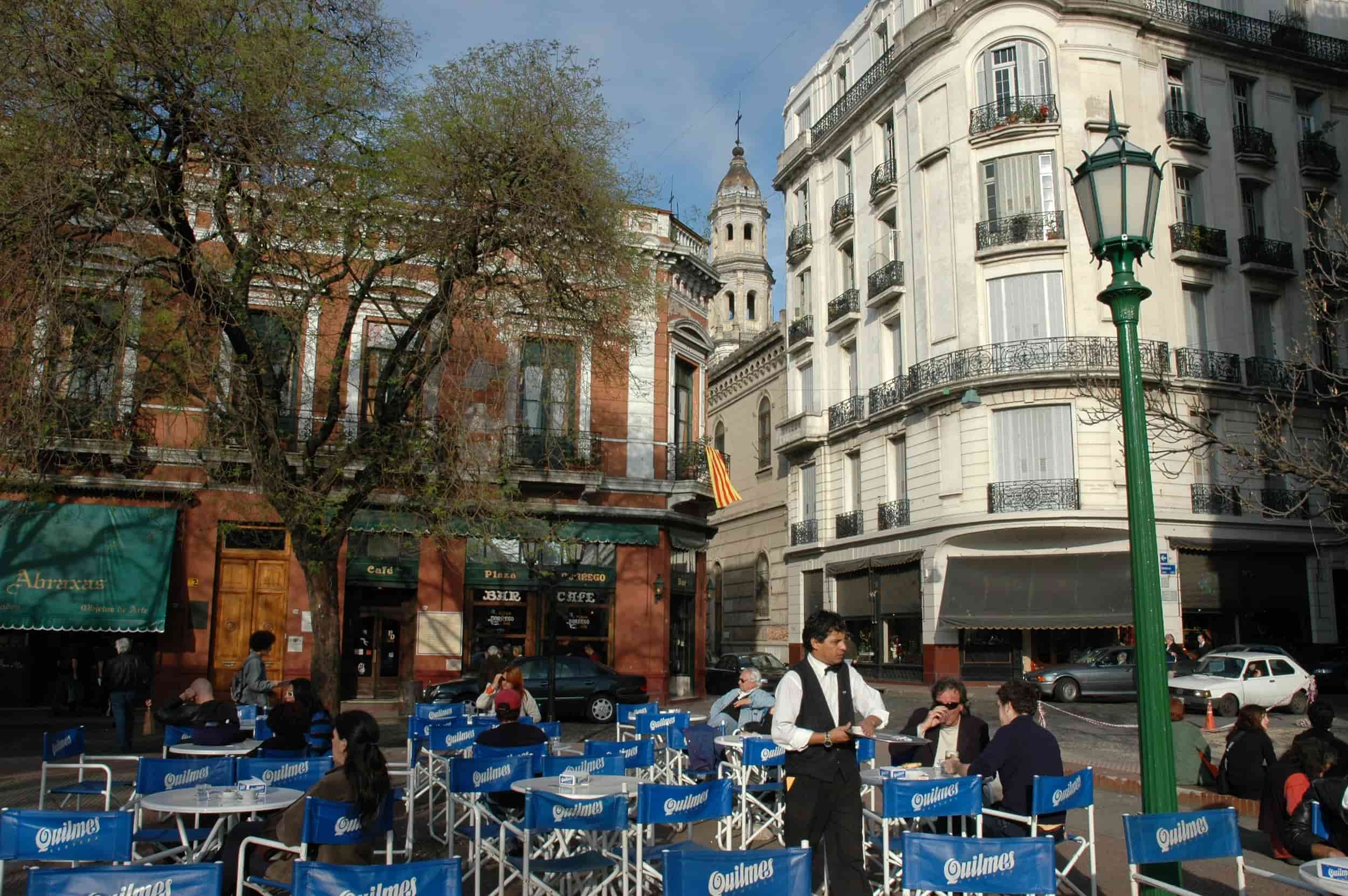 Outdoor café in San Telmo, Buenos Aires, with people sitting at blue Quilmes chairs under leafy trees, surrounded by historic colonial and European-style buildings.
