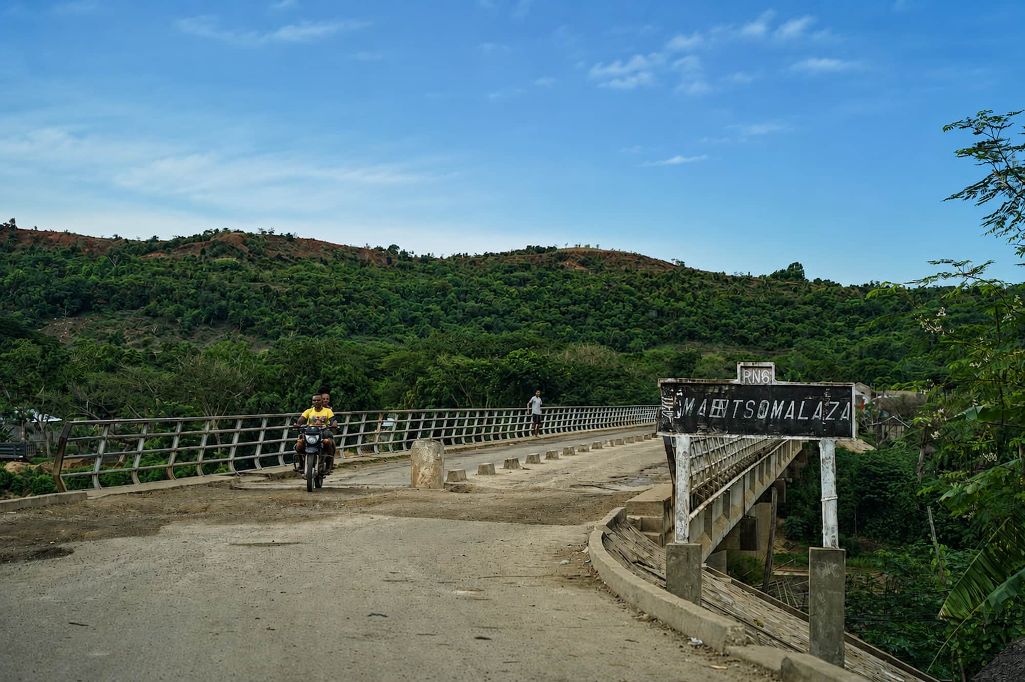 Motorcyclist crossing a steel bridge over a river on a rural road, green hills behind, Madagascar.