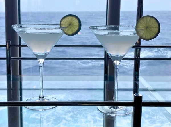 Two cocktail drinks on a glass table, with view of ocean behind them