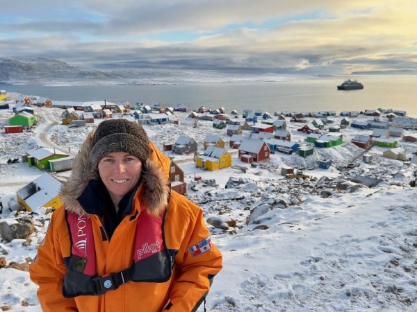 Woman in orange polar jacket, standing on a snowy hill in front of a village