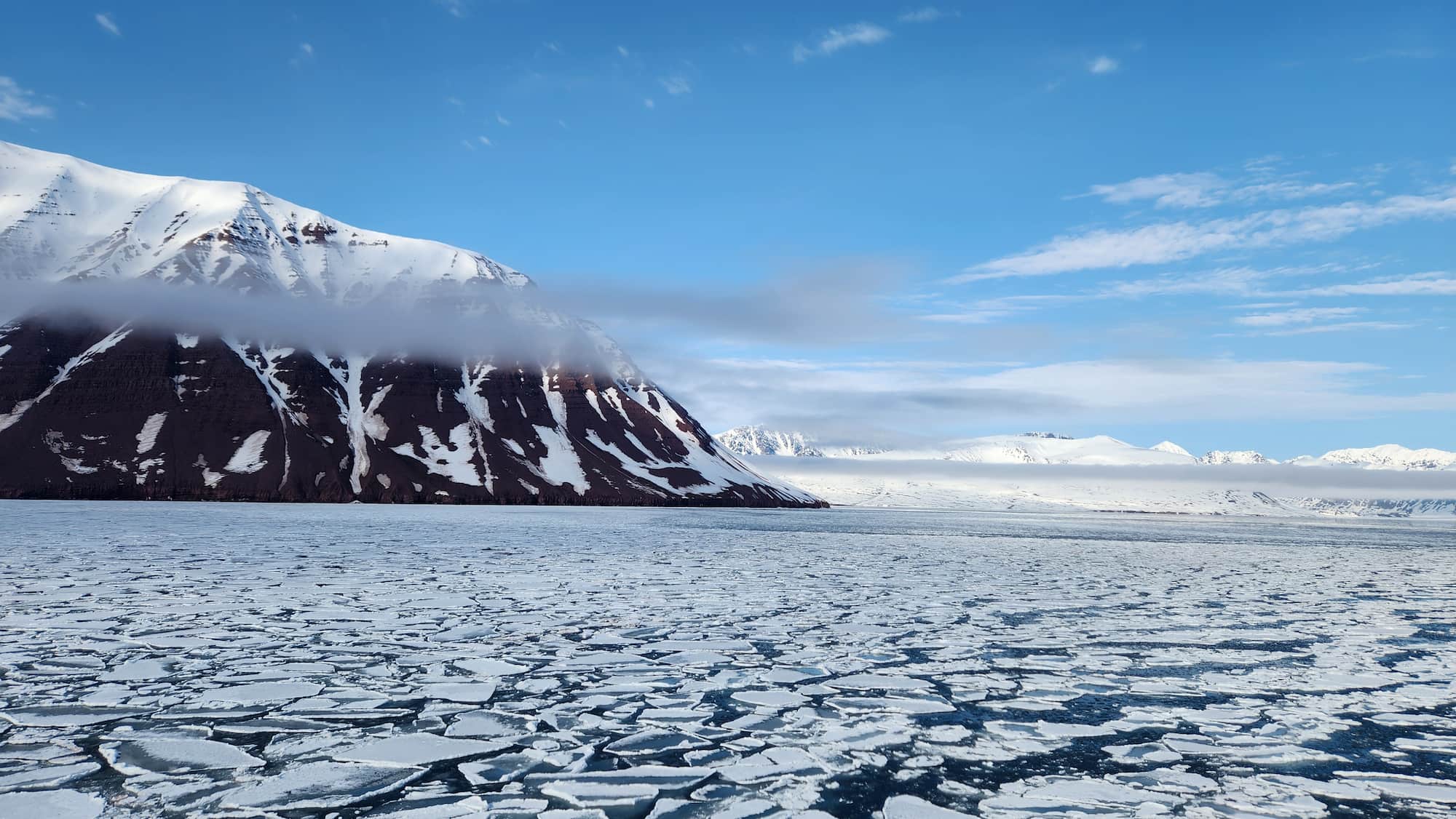 Sea ice floating beneath snow-covered Arctic mountains during an early-season expedition cruise