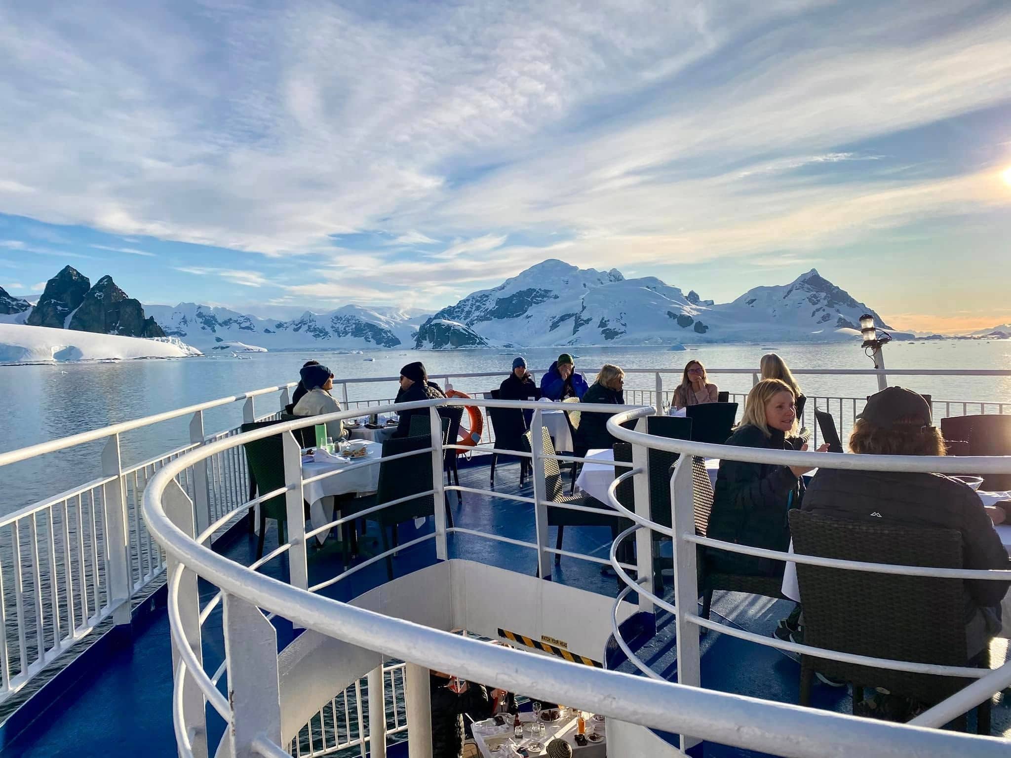 Passengers dining on the open deck of an Antarctica expedition cruise ship, surrounded by snow-capped mountains and golden evening light — November cruises offer spectacular sunset colors on the Antarctic Peninsula.