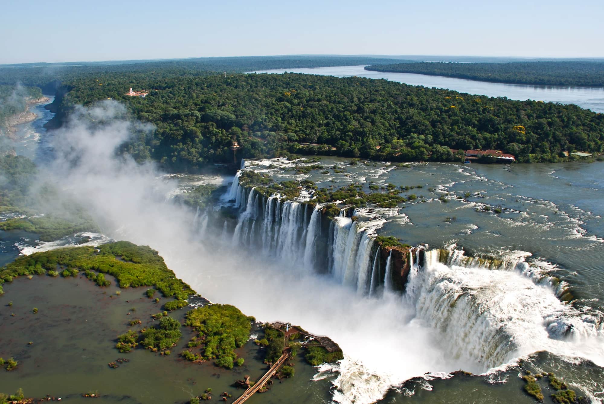 A wide aerial view of Iguazú Falls showing a long curtain of waterfalls, misty canyons, and dense green forest and walkways.