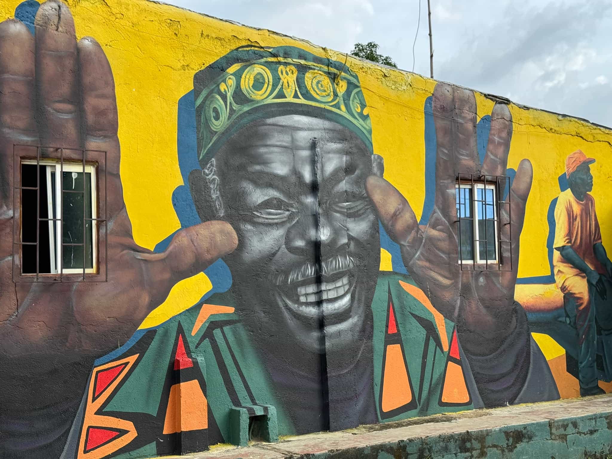 Mural of an elderly man in a patterned cap with raised hands, painted on a yellow wall in San Basilio de Palenque, Colombia.