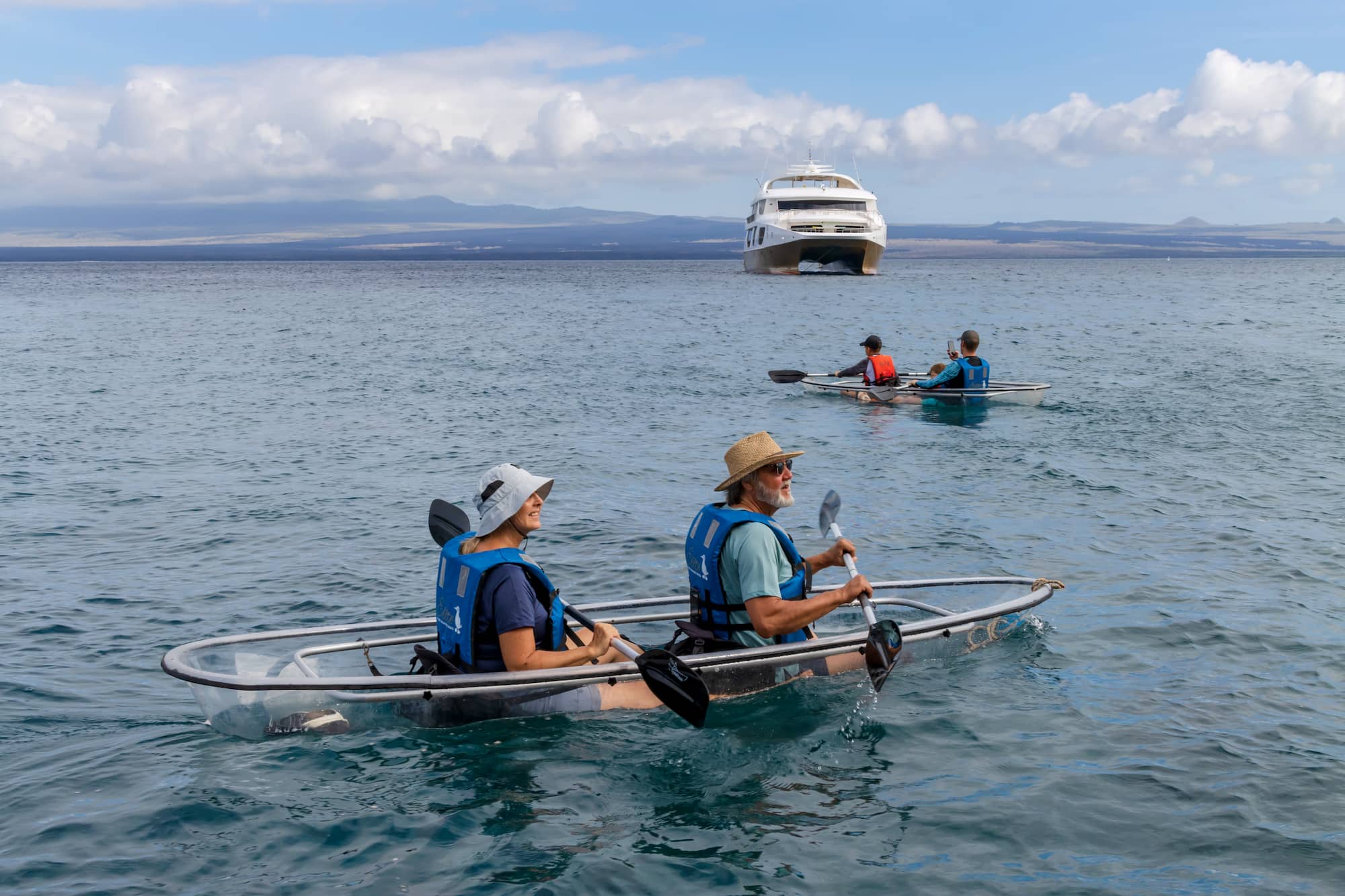 Guests paddling transparent kayaks with a small expedition catamaran anchored behind them, Galapagos Islands, Ecuador.