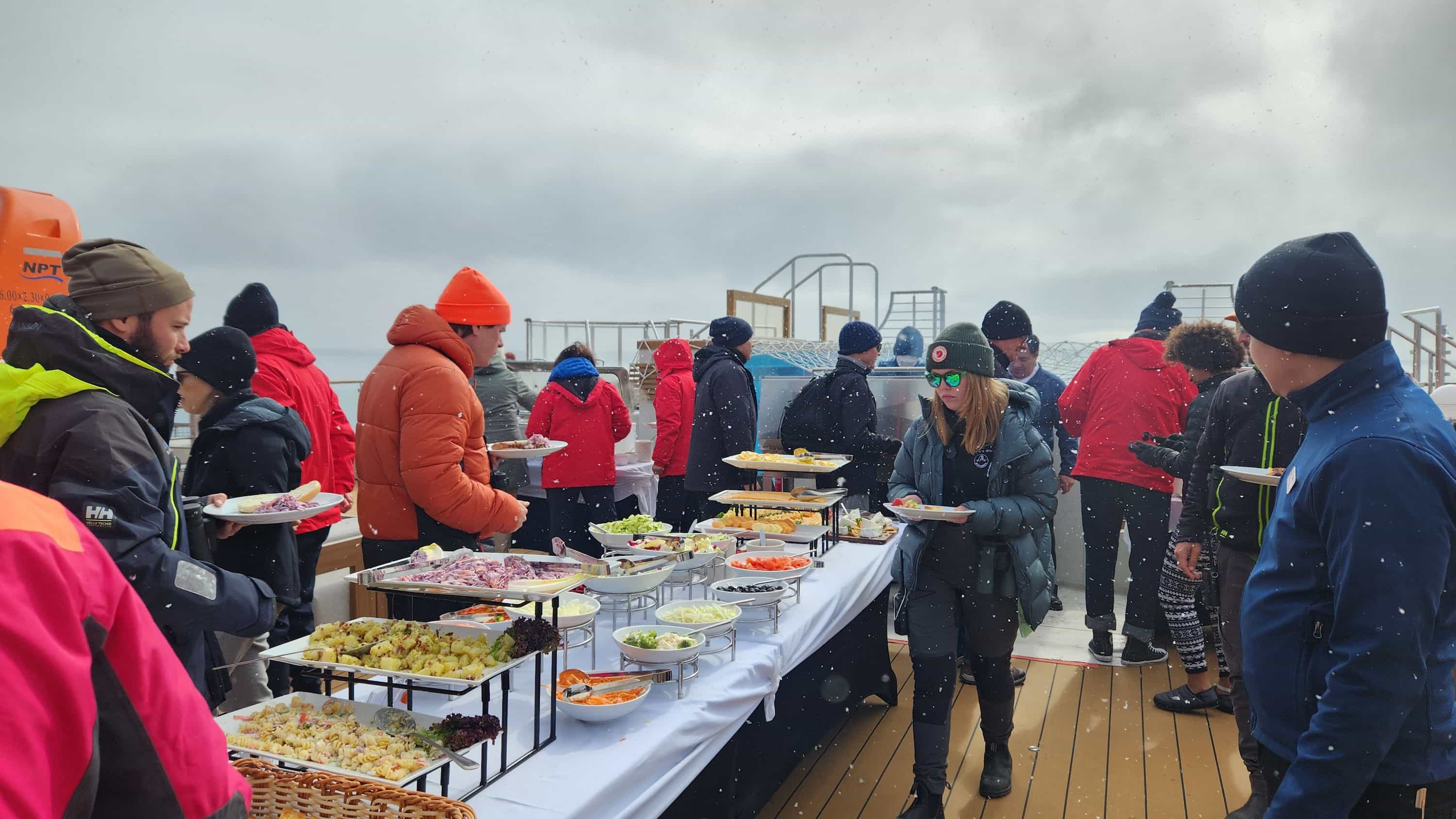 Guests dressed in warm parkas enjoying a buffet lunch on deck aboard Ultramarine, with snow falling during an Arctic cruise in Svalbard.