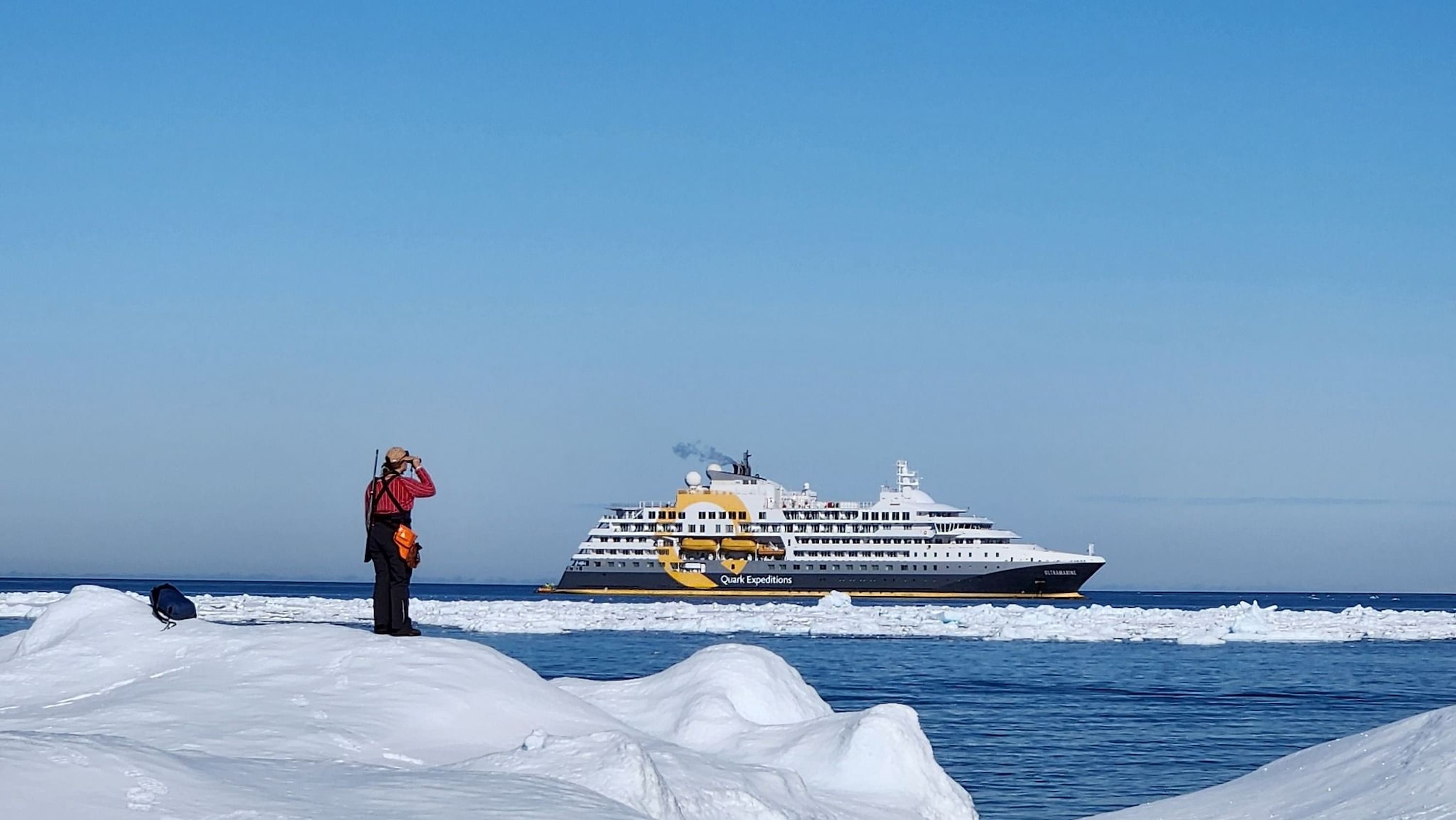 A guide stands on the Arctic ice, scanning the horizon with binoculars as the Ultramarine expedition vessel comes into view.