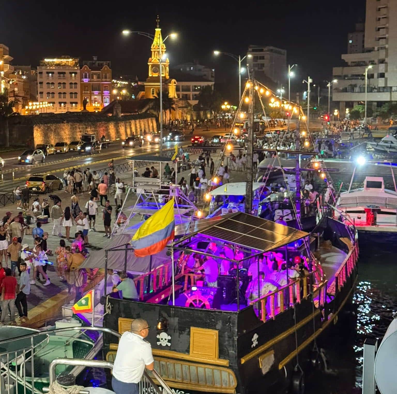 Crowds and lit-up party boats on the waterfront at night, with the Clock Tower and city walls behind, Cartagena, Colombia.