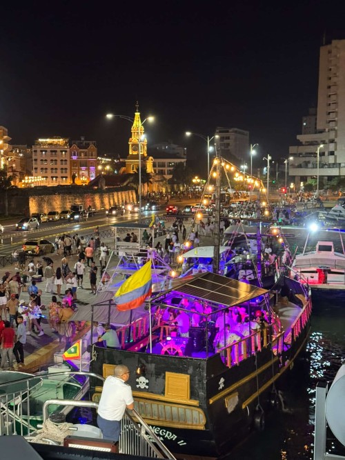 Crowds and lit-up party boats on the waterfront at night, with the Clock Tower and city walls behind, Cartagena, Colombia.