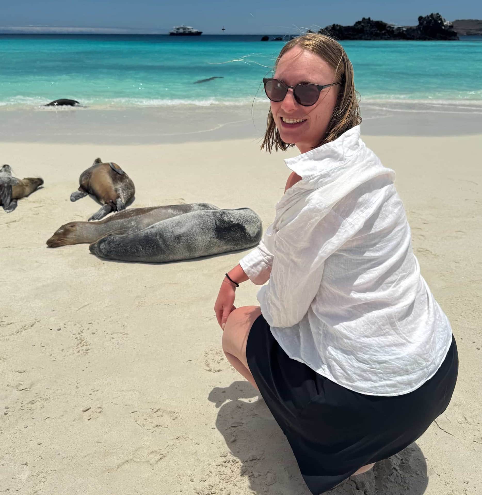 Tourists posing in front of Galapagos sea lion pups resting on a white-sand beach with a small cruise ship offshore, Galapagos Islands, Ecuador.