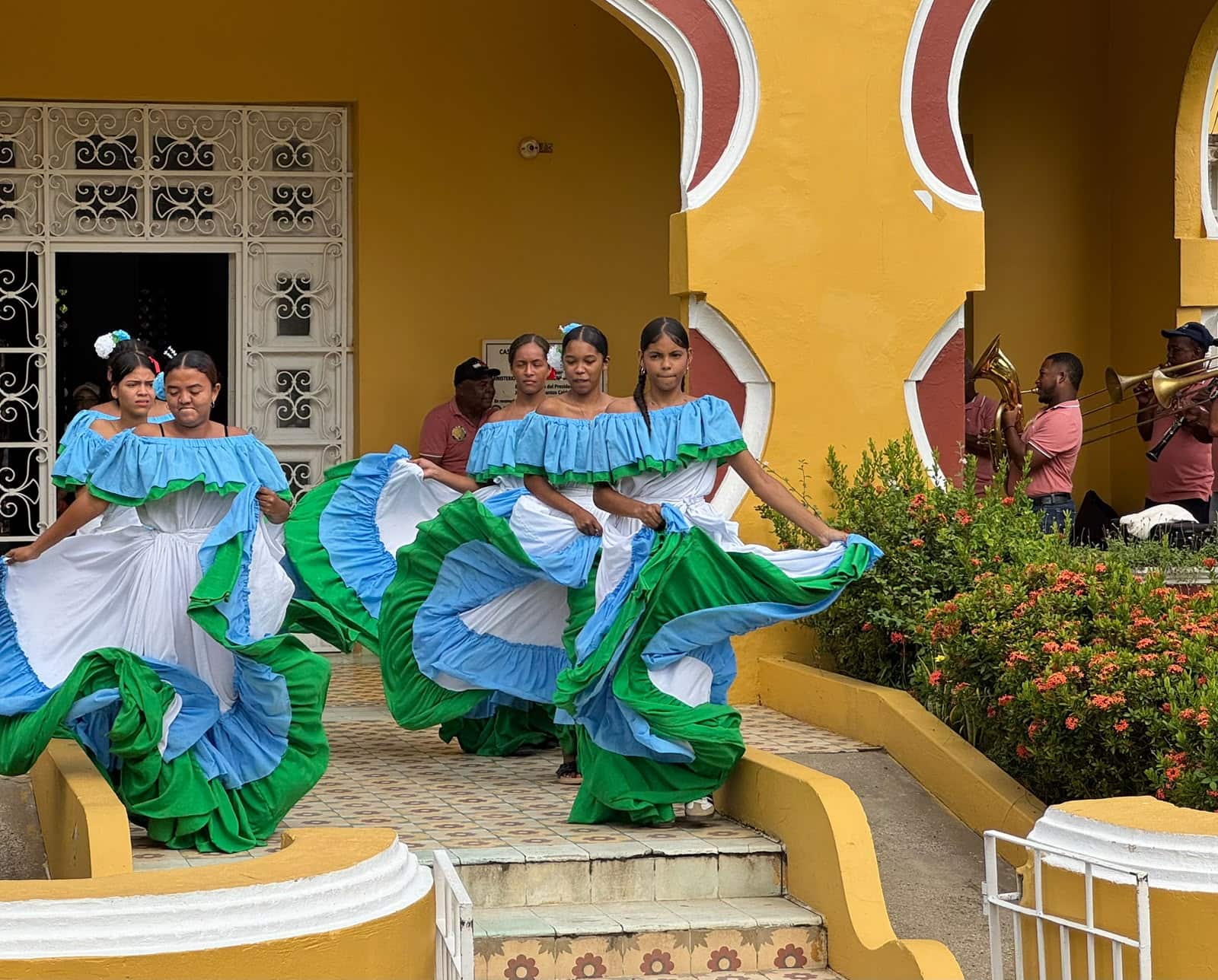 Dancers in traditional blue, white and green skirts performing to a brass band outside a yellow colonial building, Magdalena River region, Colombia.
