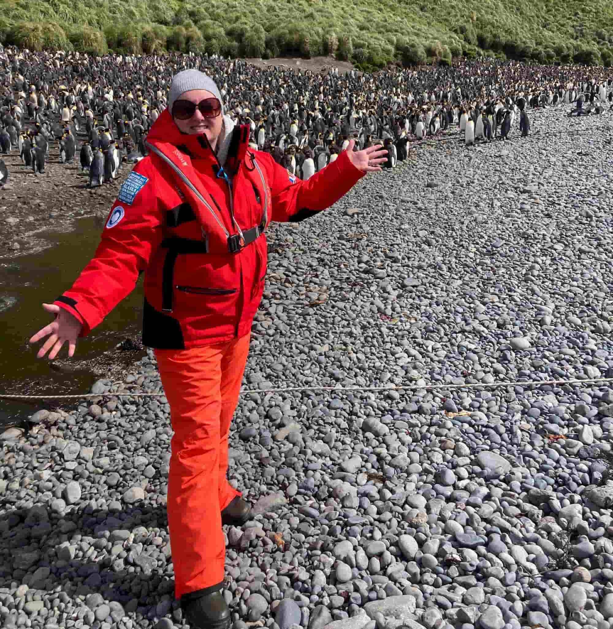 A woman landed on the sub-Antarctic island, dressed in warm, waterproof clothing, with a colony of penguins visible in the background.
